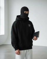 a men stand a little bit left hand side and looking front and grap a green color book and wear ninja mask hoodie with white trouser