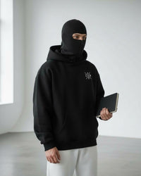 a men stand a little bit left hand side and looking front and grap a green color book and wear ninja mask hoodie with white trouser