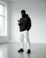 a men stand on floor wear black jogger shoes, white trouser and ninja hoodie with mask and using phone.