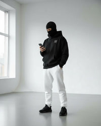 a men stand on floor wear black jogger shoes, white trouser and ninja hoodie with mask and using phone.