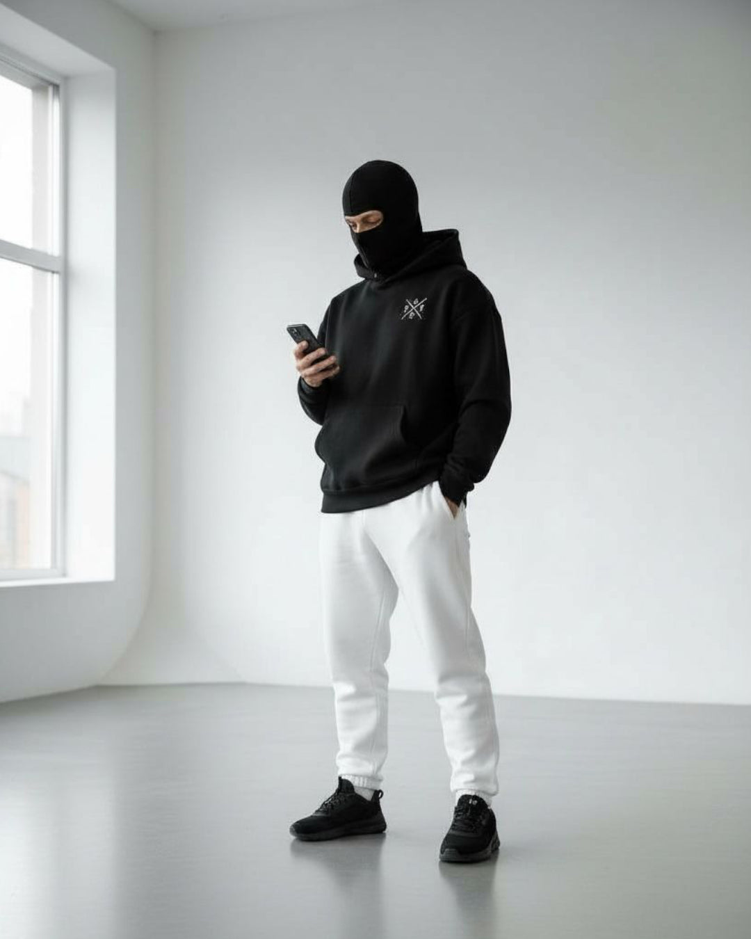 a men stand on floor wear black jogger shoes, white trouser and ninja hoodie with mask and using phone.