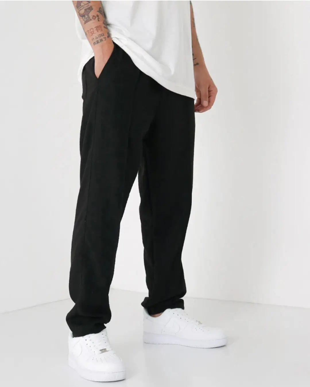 Man wearing black loose-fit pants with white sneakers and a white t-shirt, showing a relaxed casual style with visible arm tattoo